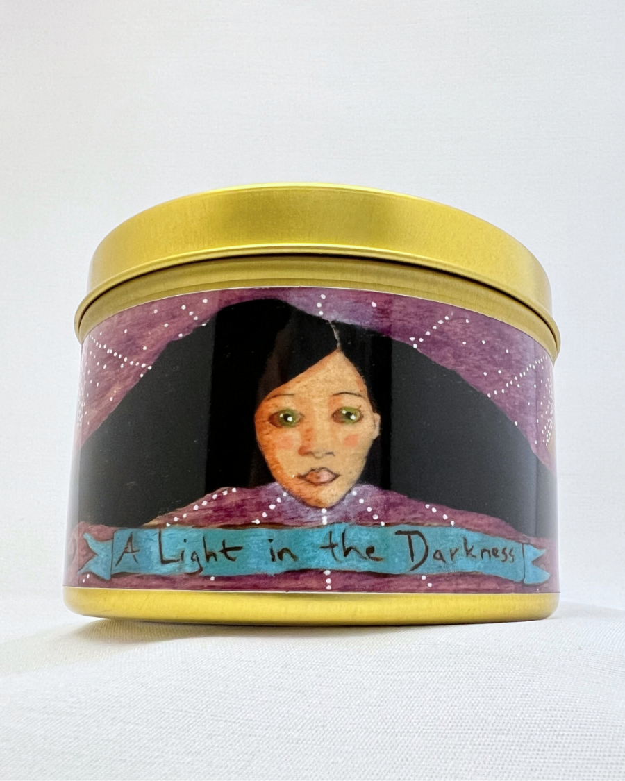 A candle in a gold metal container with art on the label. The label is purple with the face of a woman on it. She has green eyes and black hair that curls off to either side. Beneath her is a blue banner that reads "A Light in the Darkness." The art is visionary, feminine, and inspirational.