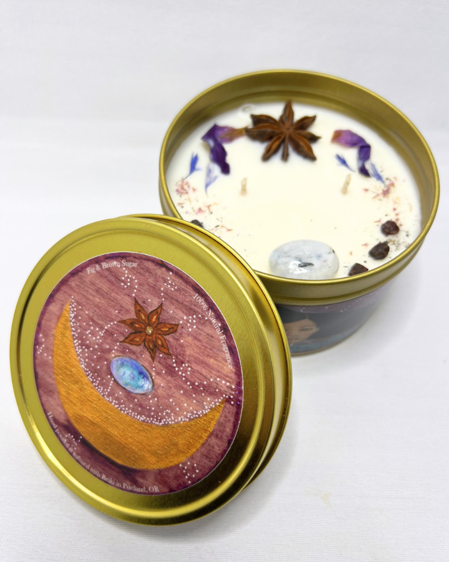 A beautiful white candle in a golden metal container. The candle has moonstone, garnet, star anise, and other decorative elements arranged in the wax. The lid has a label with art by Portland Artist Lea K. Tawd. It has a violet background with a golden crescent moon, a moonstone and star anise, with energetic white dots around them. The lid is positioned on the side of the candle, leaning up against it.