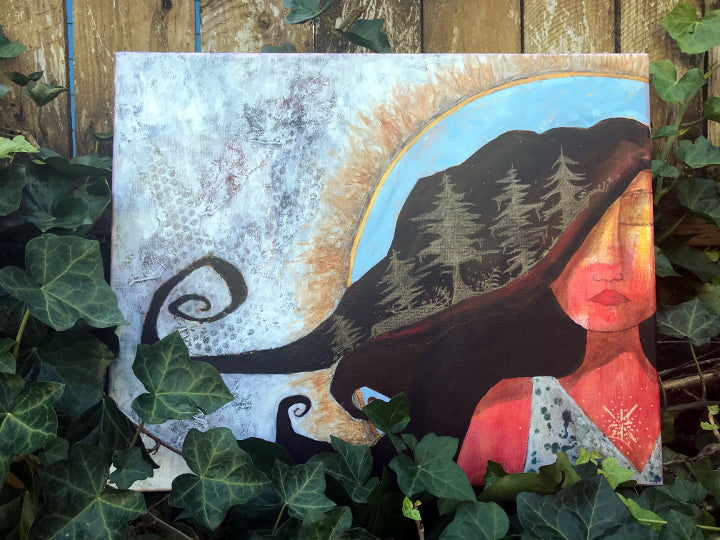 feminine artwork depicting a woman with a silhouette of a forest in her hair and organic, earth tone texture and markings behind her. the painting is sitting in some vines along a fence.
