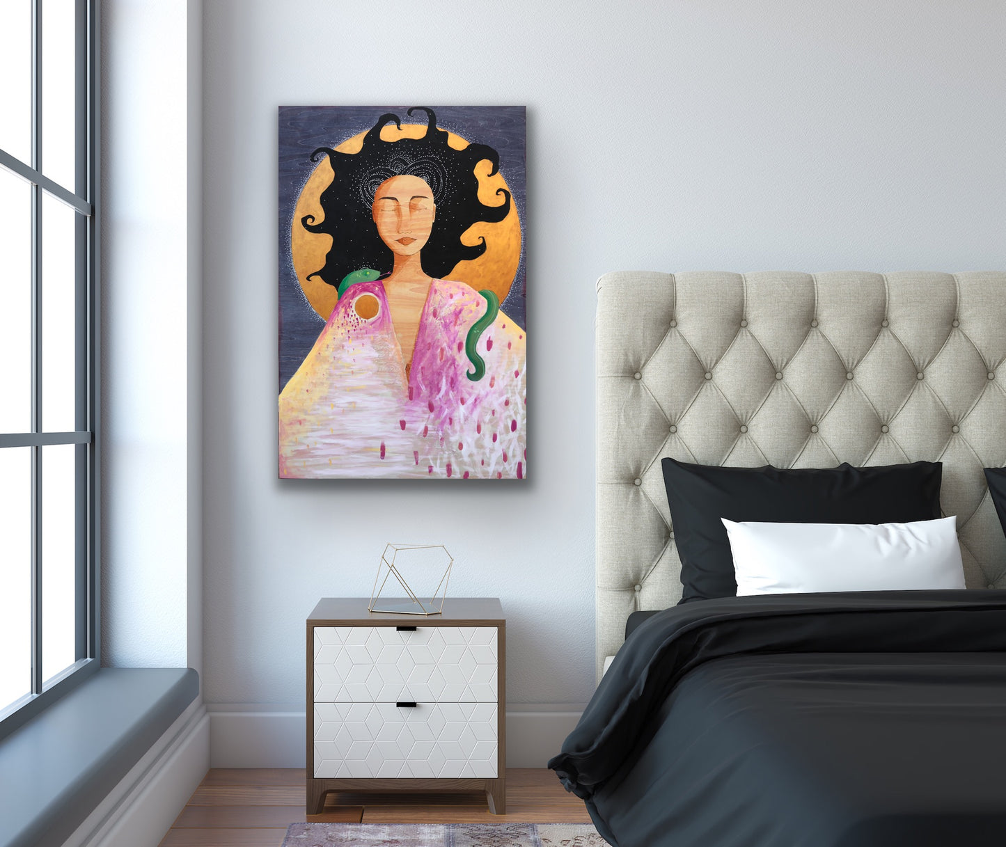 A mixed media painting of a woman with a snake draped over her shoulders is hanging on a wall over a bedside table. On the left side of the frame is a window with sunlight coming in, and on the right is a bed with black bedding and a cushioned beige headboard.