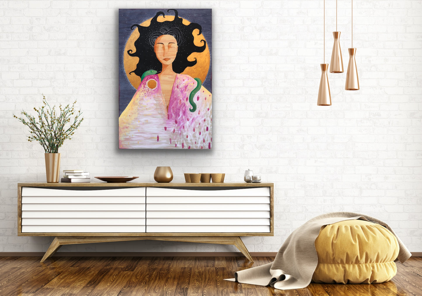 A large painting of a woman, a full moon, and a snake is hanging on a living room wall. Below the painting is a long, low cabinet with several golden or brass objects on it--cups, a vase, a plate, and some books. There are gold-colored hanging lamps to the right of the painting and a yellow pouf with a beige blanket draped over it on the hard wood floor.