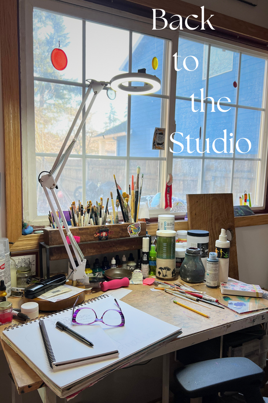 Artist's drafting table in front of a window. The table is covered in art supplies, a sketchbook, a journal, and a pair of glasses.