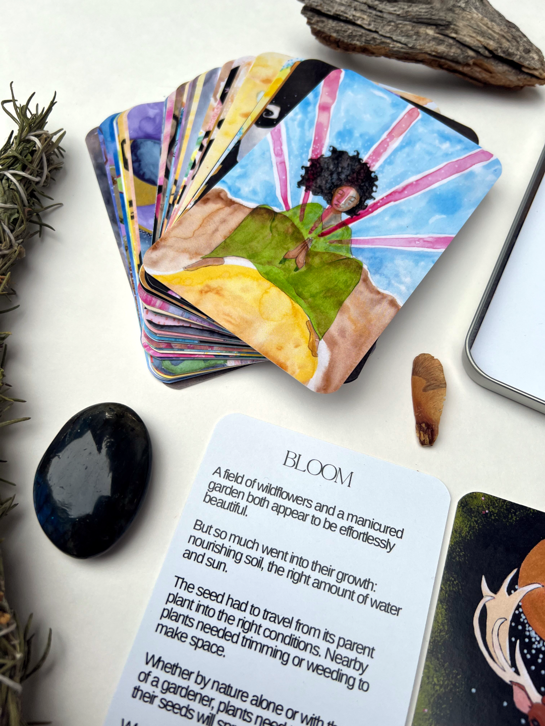 Set of oracle cards with a card titled 'Bloom' on a white surface with natural elements. The image of the card on top of the deck is a Black woman in a green gown squatting with her hands in a prayer posture and magenta beams of color emanating from her hands into the blue background.