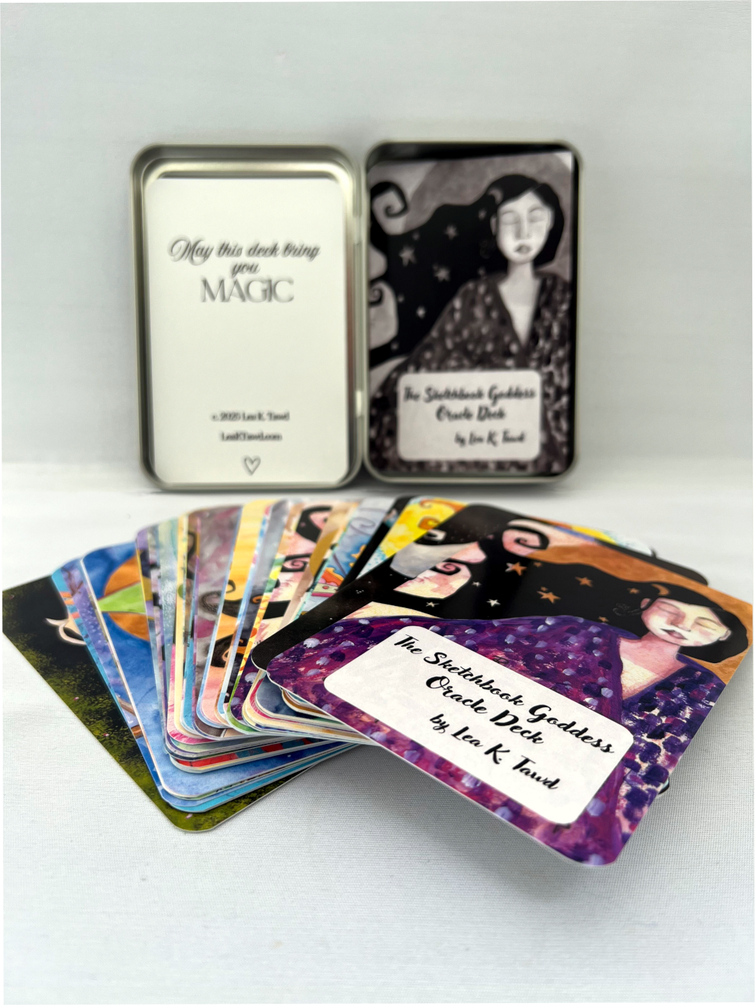 The Sketchbook Goddess Oracle Deck cards fanned out and in focus in the foreground with the tin that they come in blurred behind it. The background is a very light grey/white color.