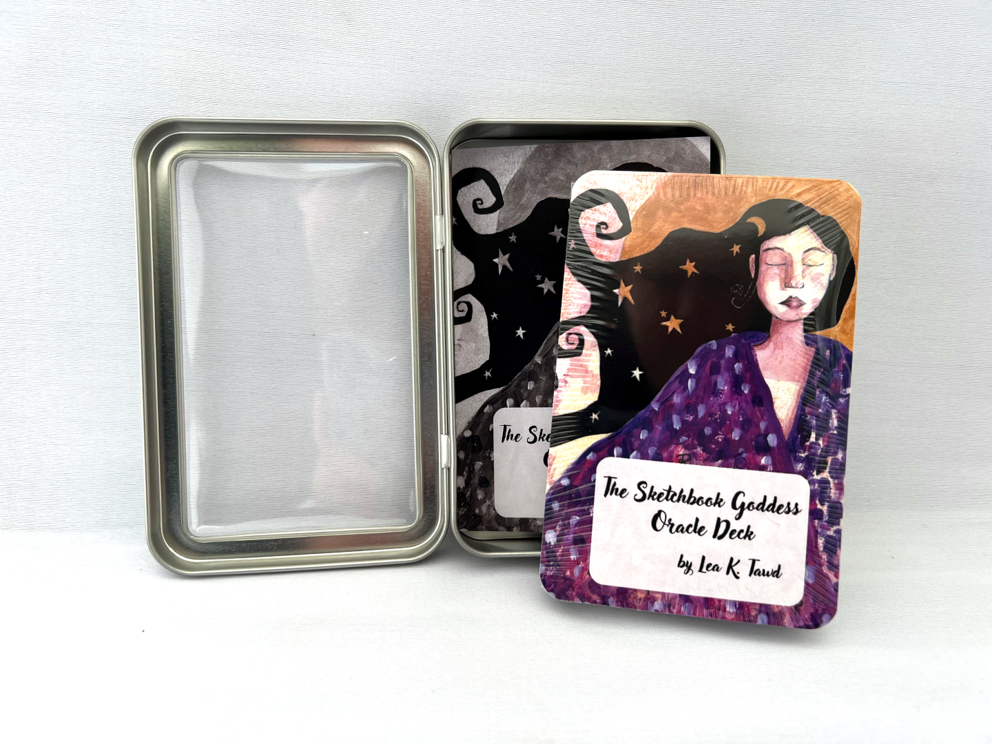 The Sketchbook Goddess Oracle Deck - The cards are shrink wrapped with the cover card showing an illustration of a white woman with her eyes closed, wearing a purple gown.  She has black flowing hair with star and moon shapes in it and gold behind her.  At the bottom of the card is a white rectangle with rounded corners and the words "The Sketchbook Goddess Oracle Deck by Lea K. Tawd." The shrink wrapped deck is leaning up against the open tin that it comes in.  The background is white.