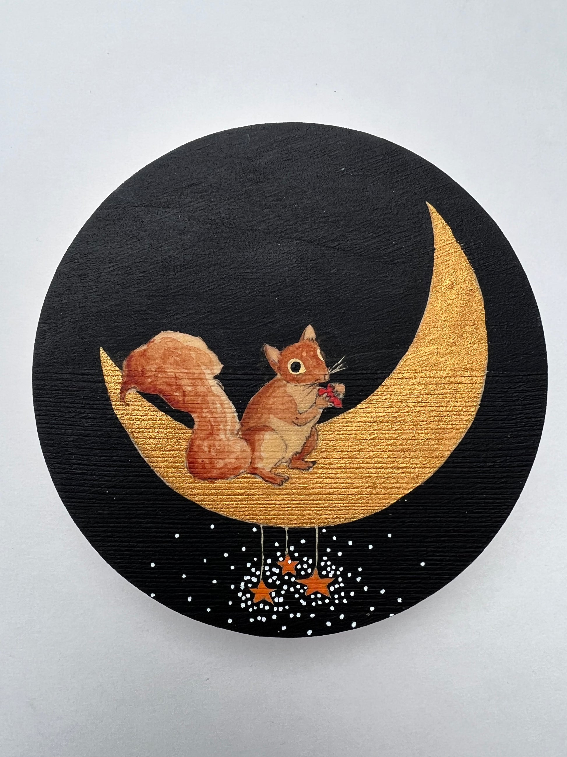 A painting of a squirrel holding a heart and standing on a crescent moon.  There are 3 golden stars hanging on strings from the moon and little dots of white light shining from the stars.  The background is black.