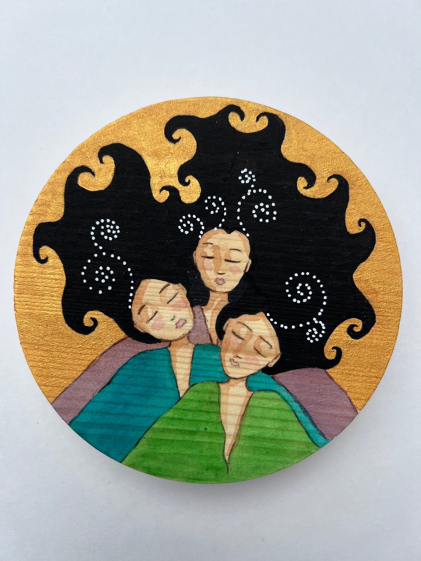 A circular painting of 3 women with black curly hair. Each wears a different top, one green, one teal and one purple.  The background is gold.