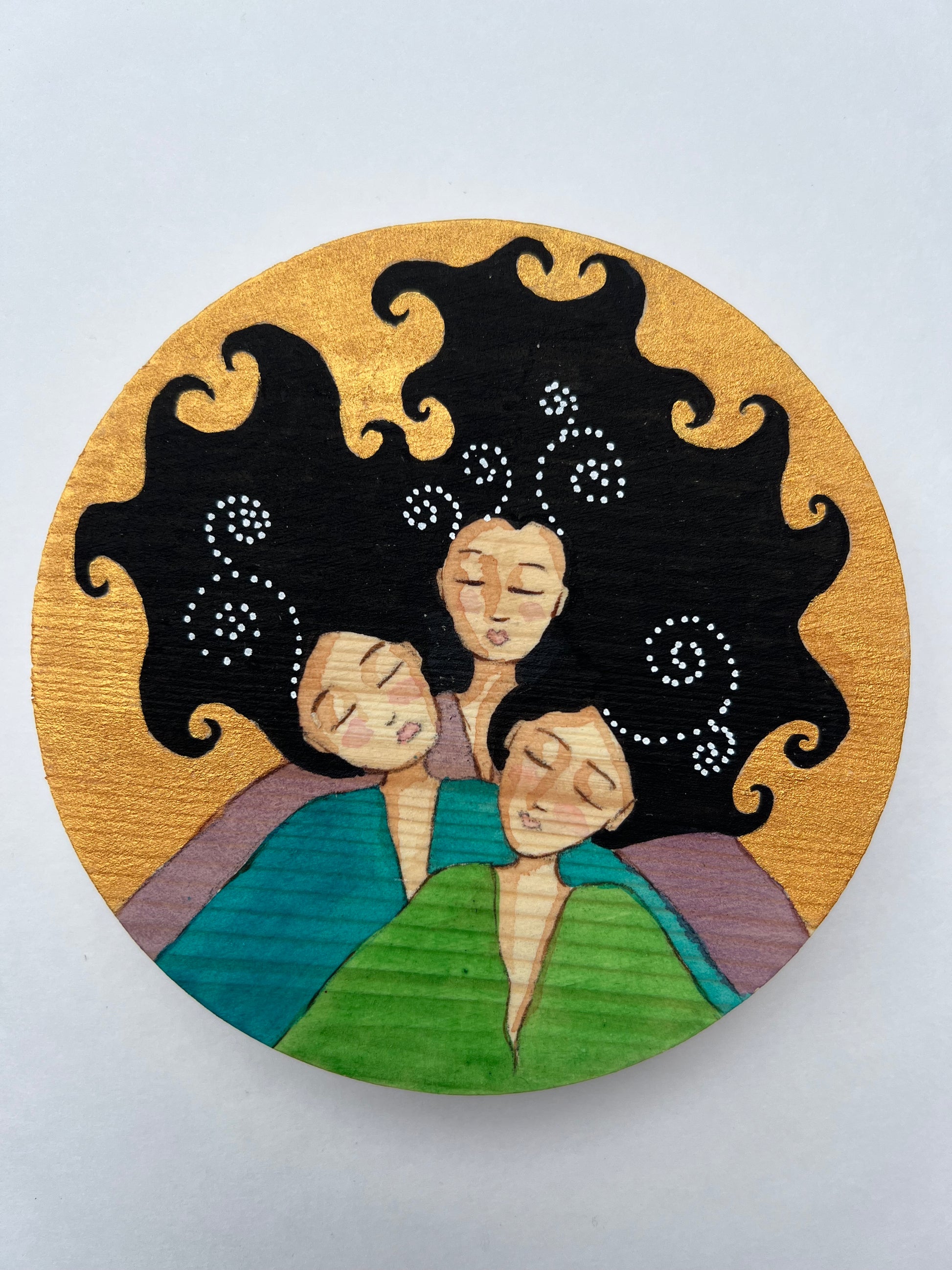 A circular painting of 3 women with black curly hair. Each wears a different top, one green, one teal and one purple.  The background is gold.