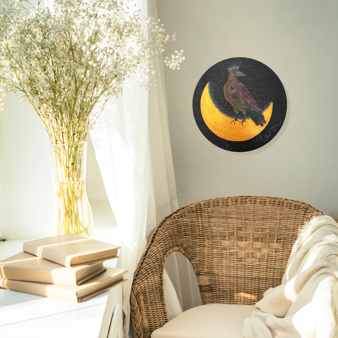 A light-filled reading nook with a wicker chair, beige-bound books, and light white flowers.  On the wall is a circular painting of a crow standing on a crescent moon.