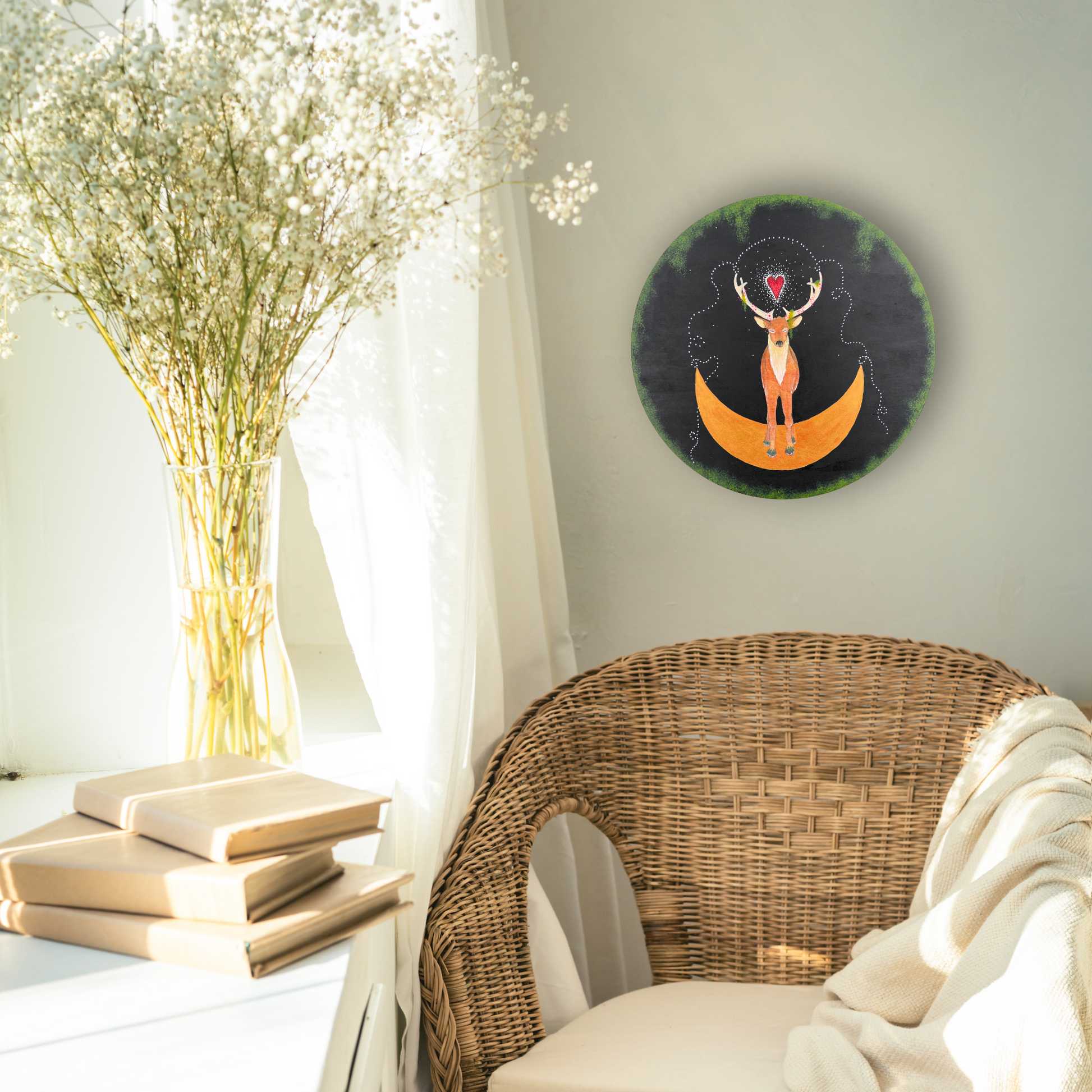 Artist Lea K. Tawd's painting, "Hart," depicting a stag standing on a crescent moon on a round substrate, hanging on the wall in the corner of a breezy, well-lit room.  There are white curtains, some white flowers in a vase, three neutral-colored books on a white shelf, and a wicker chair with white cushions in the room. 