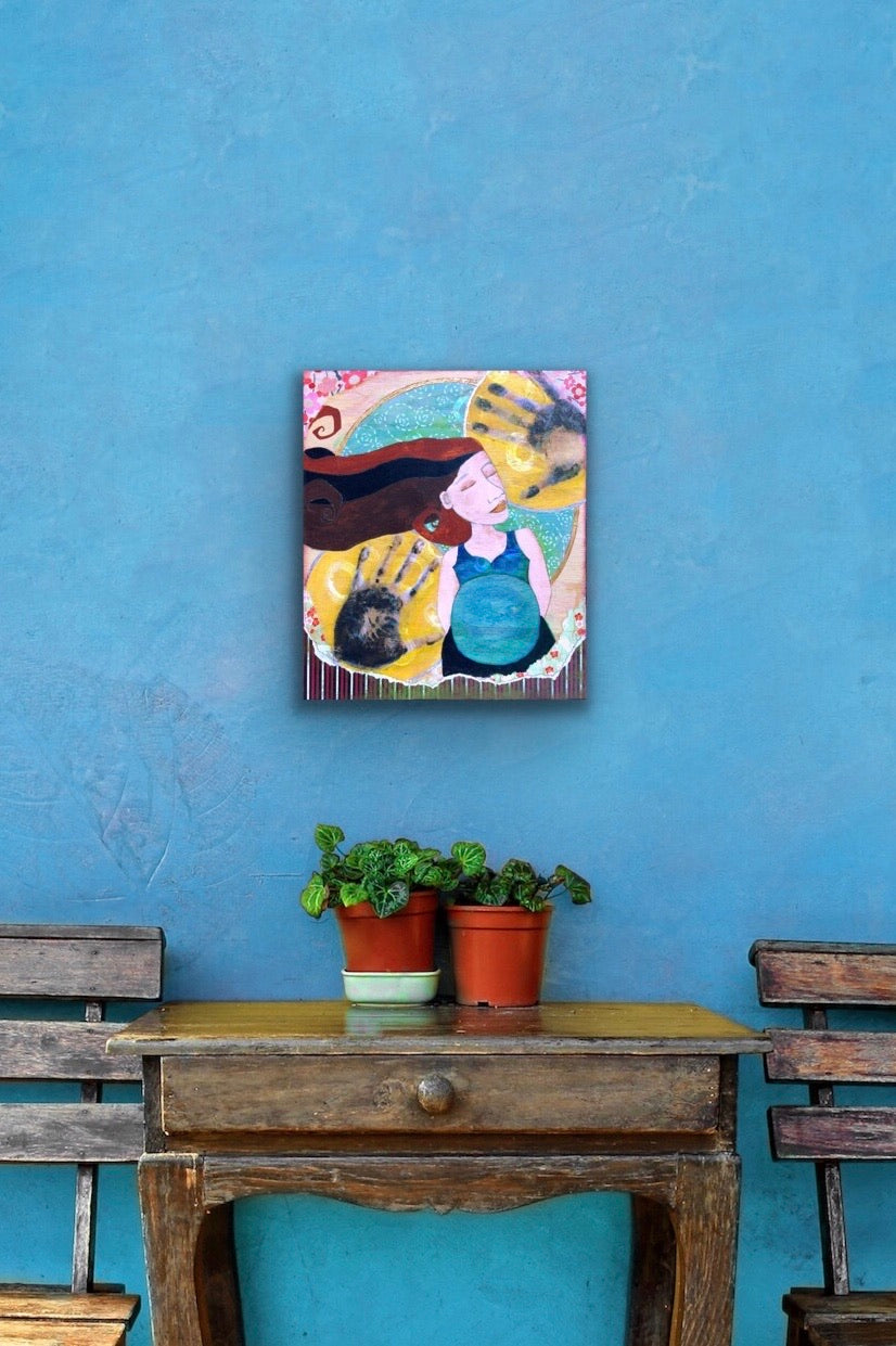 mixed media feminine painting of pregnant woman with dark hair and a blue shirt hanging on a blue wall above a small wooden table and chairs